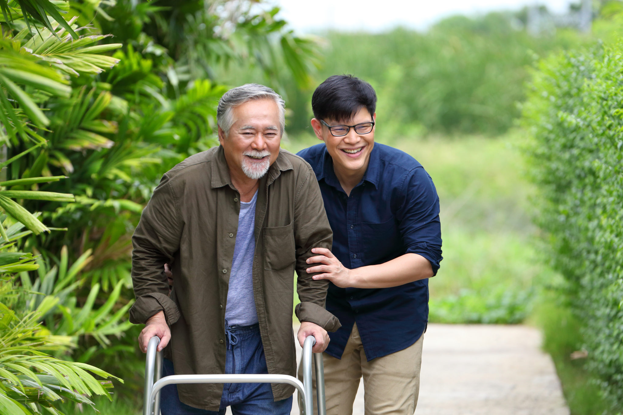 Happy elder Asian man using walker while walking for exercise around the garden with his son taking care of him at the retirement age with copy space