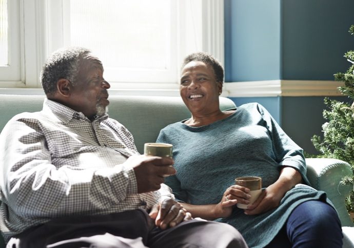 Smiling senior couple holding drinks talking while sitting on sofa at home
